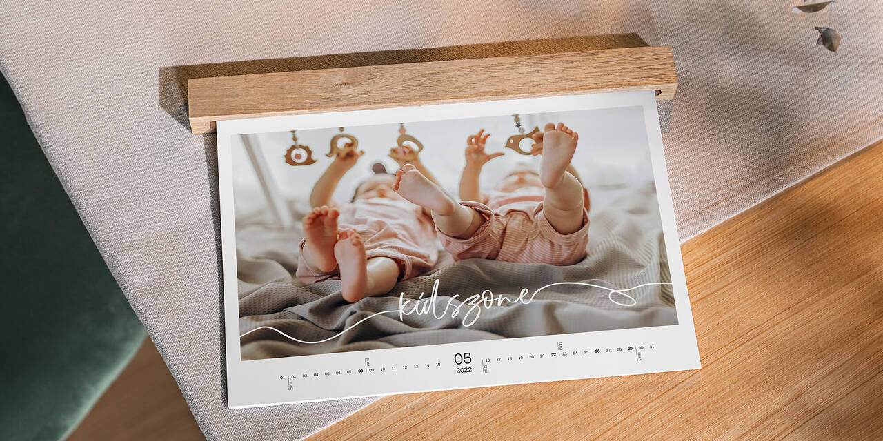 Auf einem Tisch aus Holz liegt ein Wandkalender mit Babymotiven im Querformat. Sichtbar ist das Blatt für den Monat Mai. Eine Holzleiste verdeckt die Ringbindung des Kalenders.