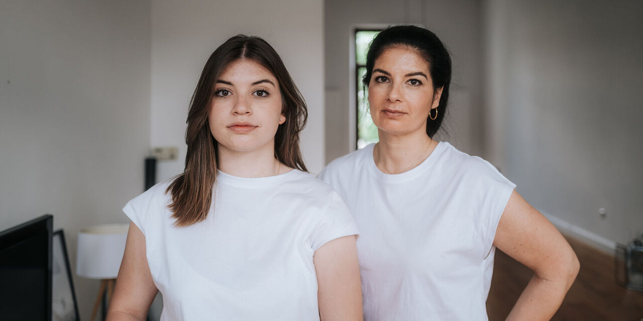 Zwei Frauen mit dunklen Haaren und weißen T-Shirts stehen nebeneinander und schauen in die Kamera.