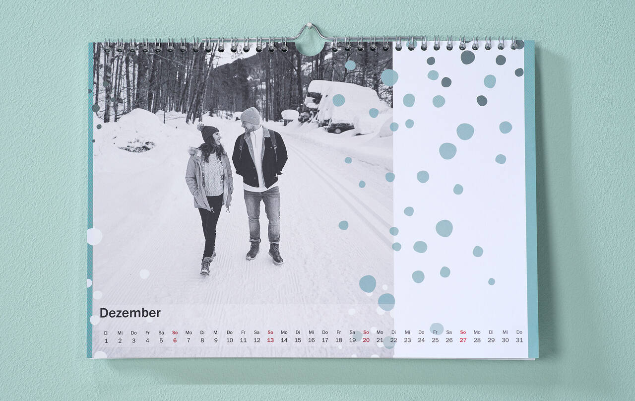 Kalender mit winterlichem Pärchenfoto und Pünktchen-Cliparts hängt an Wand.