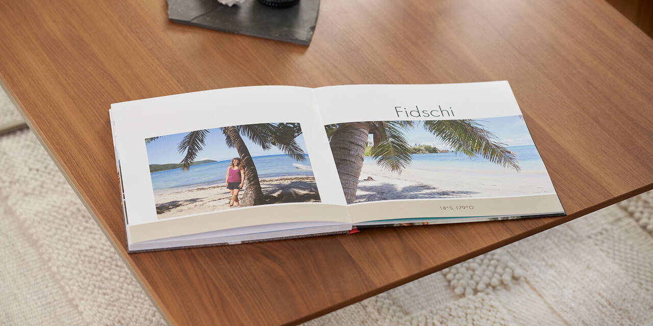 Auf einem Holztisch liegt ein aufgeschlagenes Fotobuch mit zwei Strandbildern und der Überschrift Fidschi. Über dem Fotobuch steht ein dunkles Tablett mit einer Kerze und Muschel. Unter dem Tisch sieht man einen hellen Teppich.